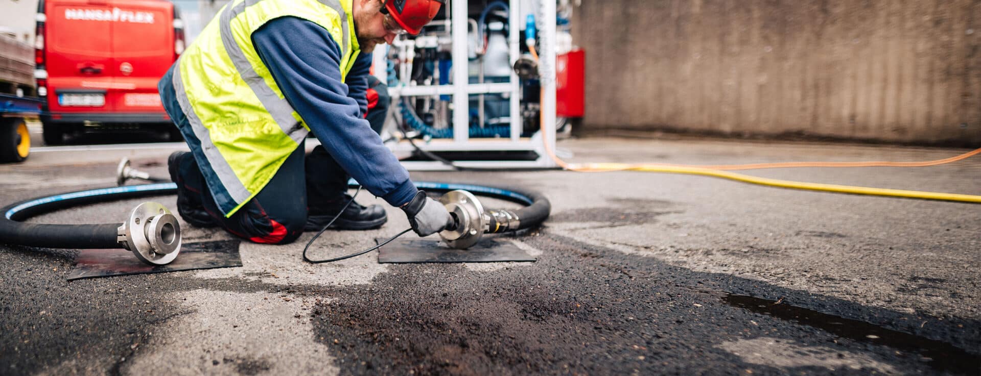 DGUV-Prüfung von Hydraulikschlauchleitungen. Mann mit Warnweste prüft Schlauch auf Boden.